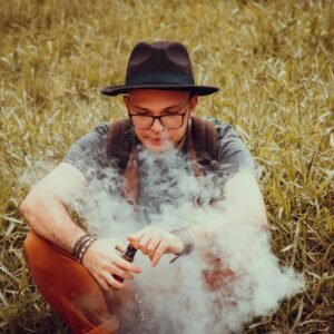 A man with a cowboy hat is sitting in a grass field, vaping on a clear day.